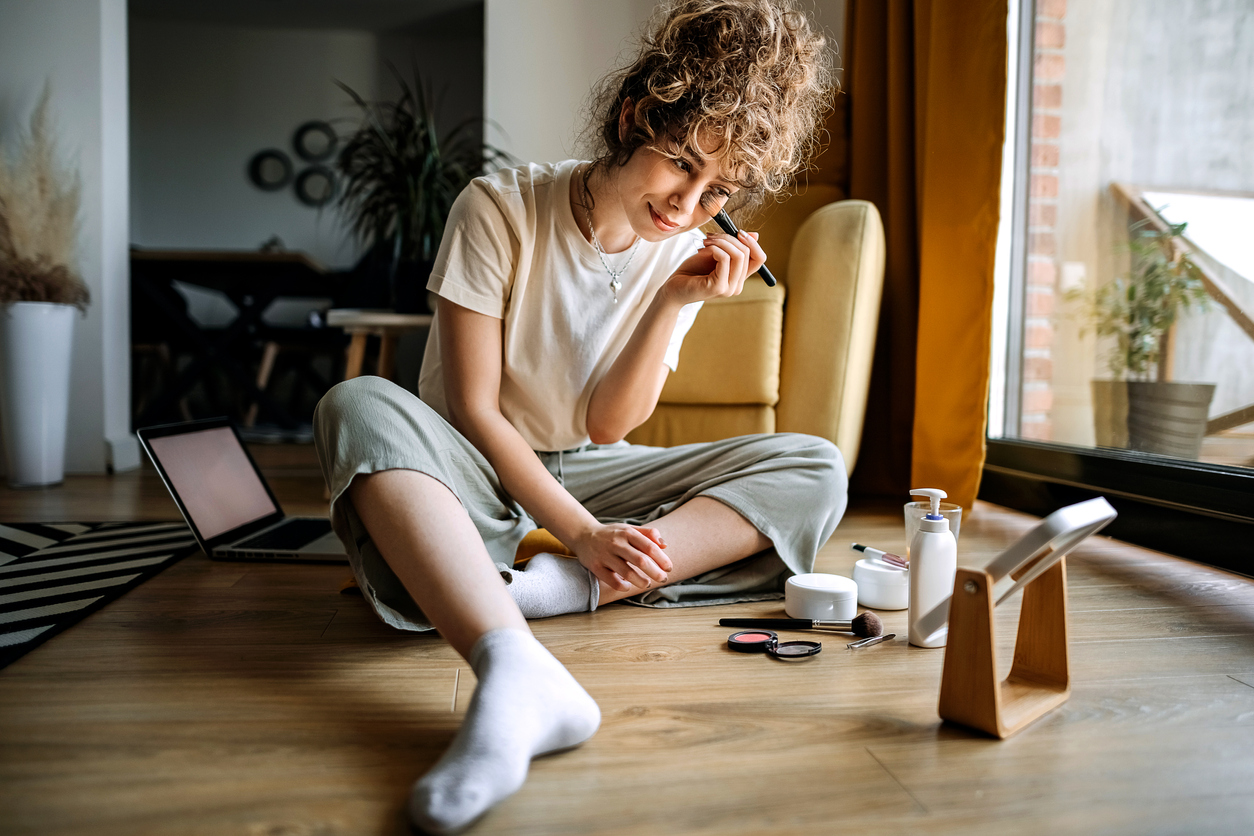 Young beautiful woman is sitting in her living room and putting on make-up. Glossier failed to evolve as a beauty brand.